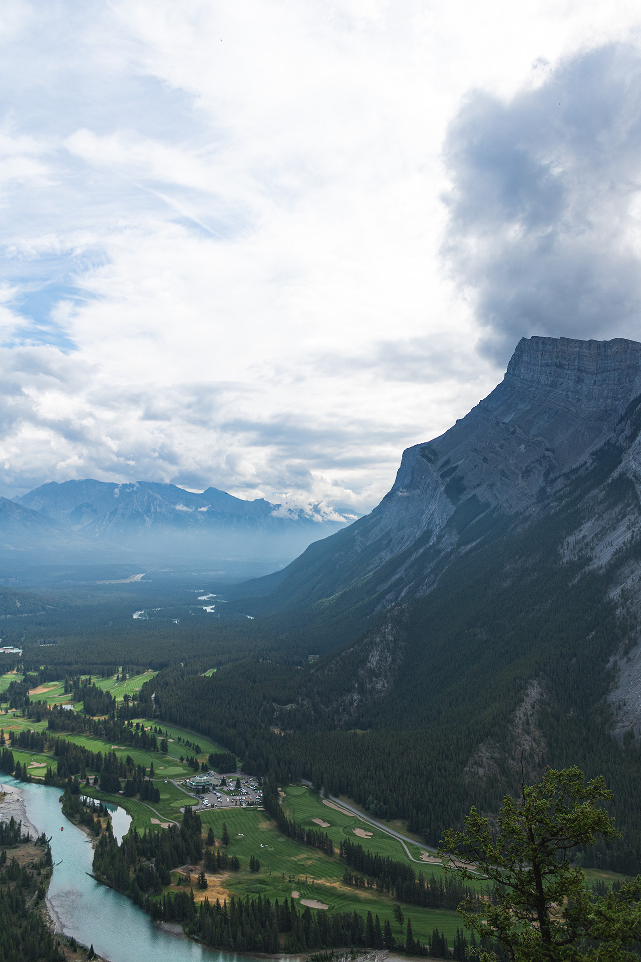 Valley with a golf course between a tall rocky mountain and a river running parallel to it, seen from halfway up the mountain's height.